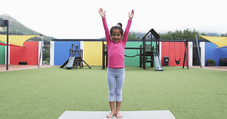 In a school playground, a young African American girl practices yoga. She has a joyful expression, wearing a pink top and gray leggings on a yoga mat, unaltered, slow motion