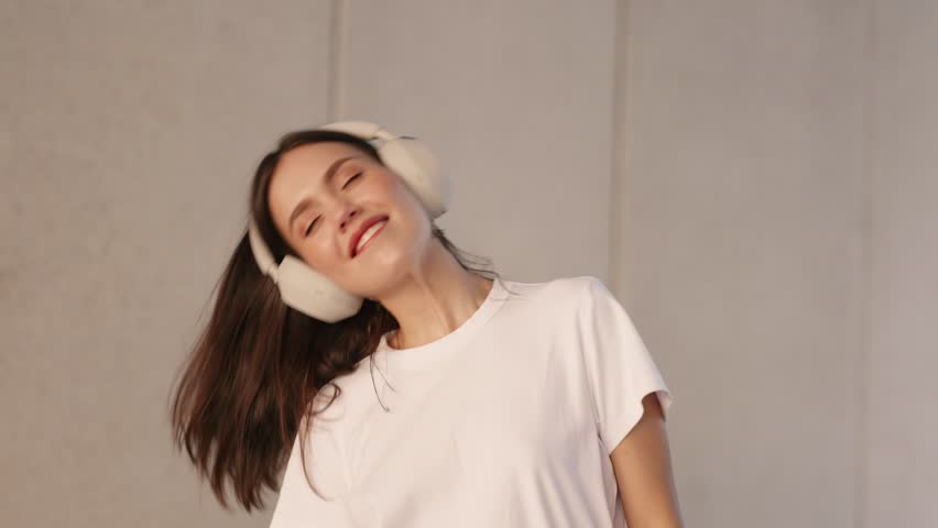 A happy teenage girl dances in a white t-shirt, enjoying music through her headphones with a joyful expression.