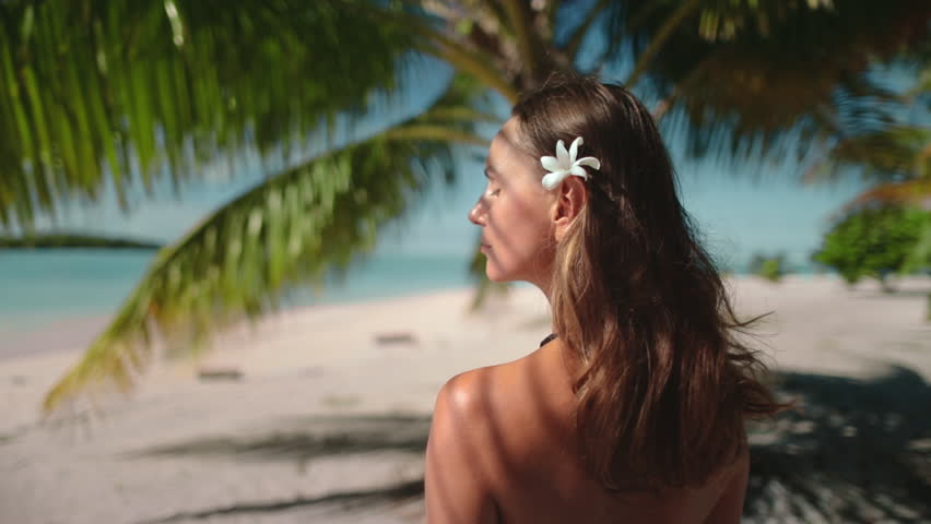 A woman is standing on a sandy beach next to a tall palm tree, under the bright sun, with the ocean in the background.