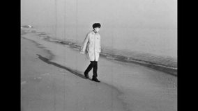 Smiling teenage boy walk along sea sandy beach in winter. Lonely young man in coat, hat run away from cold ocean waves. Seaside life outdoors. Calm waves. Vintage black white film. Retro archive 1980s