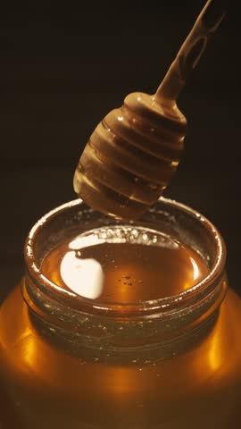 Healthy organic honey in a jar. Transparent glass jar with sweet golden honey sparkling in the sun, honey dipper inside jar on the wooden table, close up. Vertical video.