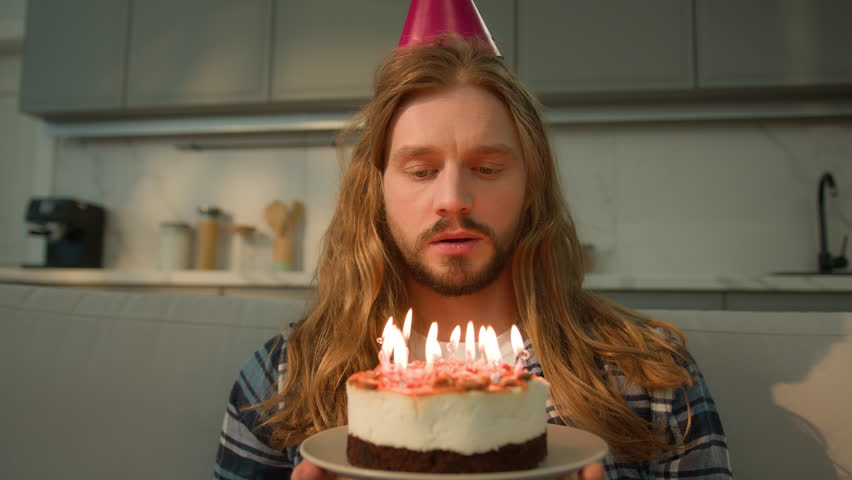 Alone sad upset frustrated one Caucasian man guy male at home apartment holding birthday cake blowing candles blow wish stress depressed lonely celebration festive hat despair disappointed immigration