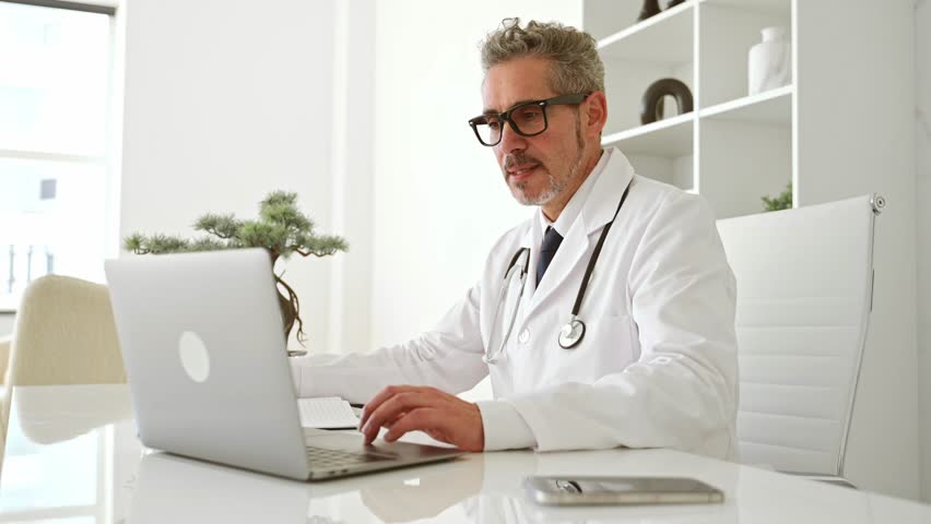 A genial mature doctor in a white coat waves hello into laptop during online appointment, seated in a well-organized clinic office that reflects a clean and efficient medical environment, taking notes