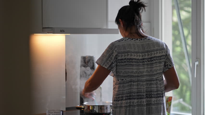 Woman Prepares Meal On Induction Stove - Stainless Steel Pot Emitting Steam As She Stirs, Natural Light From Apartment Window. Casual Domestic Everyday Candid Scene