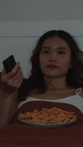 Vertical shot of young Asian woman resting in bed and eating snacks while watching TV at night alone