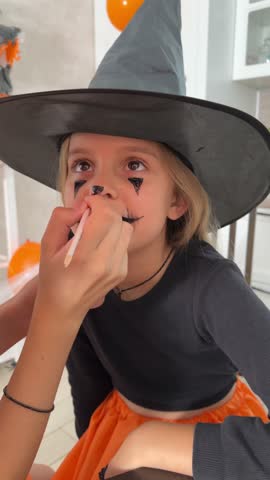 a little girl in a Halloween costume applies makeup for the holiday, a girl in a hat and witch costume paints her face for Halloween celebration