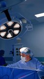 Male Caucasian mid-aged surgeon performs surgery sitting near the patient. Doctor works under the round lamp. Vertical video.