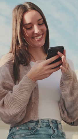 VERTICAL VIDEO, Smiling girl student sits next to the building of the university, uses a mobile phone