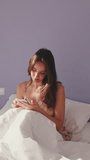 VERTICAL VIDEO, Happy woman texting on phone after waking up in the morning. Smiling girl having an online conversation, replying tap message while sitting in bed at home