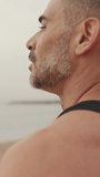 Vertical video, Close-up of middle-aged athletic man standing on the beach after workout and looking at the seascape