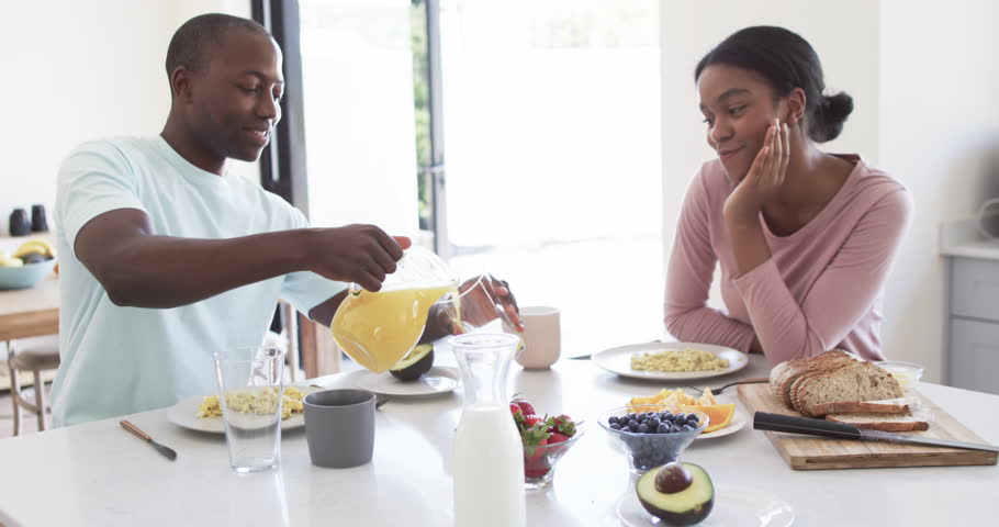 African American couple enjoys breakfast together at home in the kitchen. The husband pours juice while the wife, with a ponytail, watches with a smile, unaltered, slow motion. Royalty-Free Stock Footage #3476888259