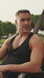 Vertical video, Middle aged muscular man in sportswear relaxing after workout while sitting in park outdoors