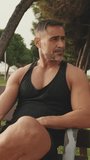 Vertical video, Middle aged muscular man relaxing after workout while sitting in park outdoors