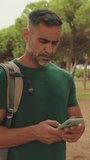 Vertical video, Mature man wearing casual clothes, with backpack on his shoulder, stands in the park, sends SMS from mobile phone, always online,Sideways movement