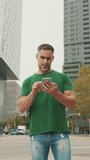 Vertical video, Mature male tourist wearing casual clothes, stands on modern buildings background, looks around, uses navigator on smartphone, uses map app