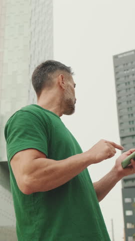 Vertical video, Mature male tourist wearing casual clothes, standing on modern buildings background, looking for right direction on smartphone, using map app, bottom view