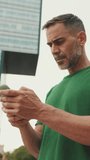 Vertical video, Mature man wearing casual clothes, standing on modern buildings background, texting on smartphone