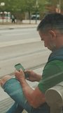 Vertical video, Middle-aged man dressed in casual clothes, uses mobile phone while waiting for tram at public transport stop in the city, view through the glass