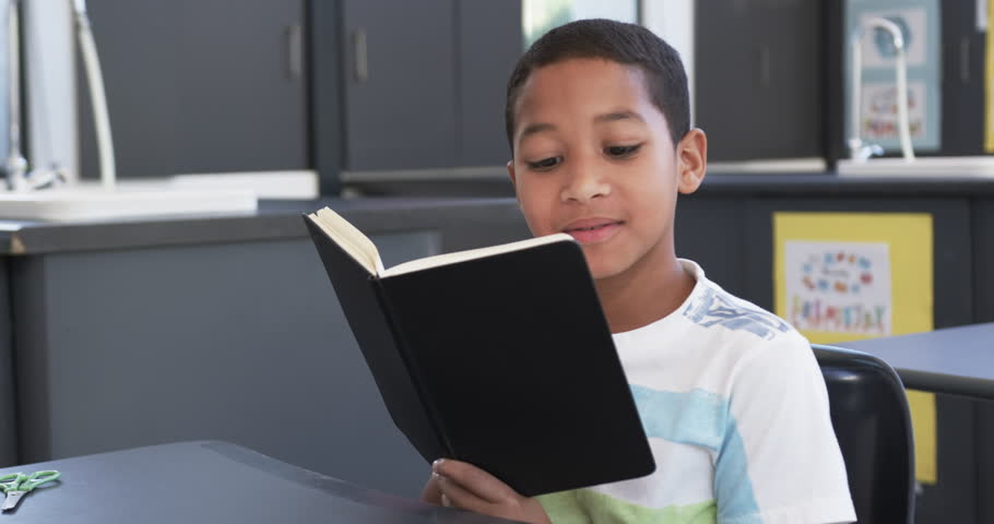 In school, in the classroom, a young African American student reads a book. He has short black hair, a focused expression, and is wearing a casual striped shirt, unaltered, slow motion. Royalty-Free Stock Footage #3476917641