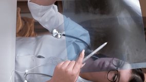 Vertical video. Doctor telehealth. Online diagnosis. Medical consultation. Woman physician examining lung x-ray film explaining to patient results on call clinic interior.
