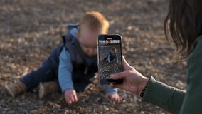in the playground, one year old red hair  boy sits on the ground and plays with the ground, explores nature, the mother takes photos and videos on the phone for childhood memories