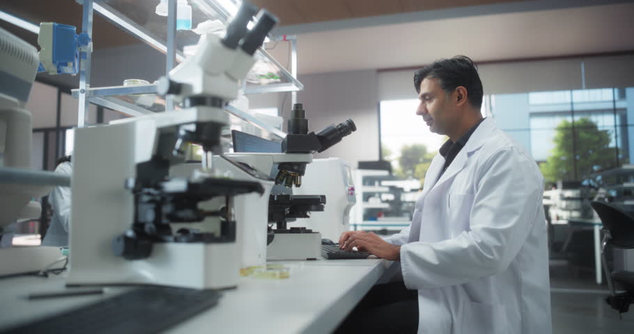 Cinematic Zoom In Portrait of an Indian Scientist Working in a Laboratory, Using Advanced Microscope, Writing Down Research Data on a Computer. Multiethnic Biotechnology Specialist Using Modern Tools