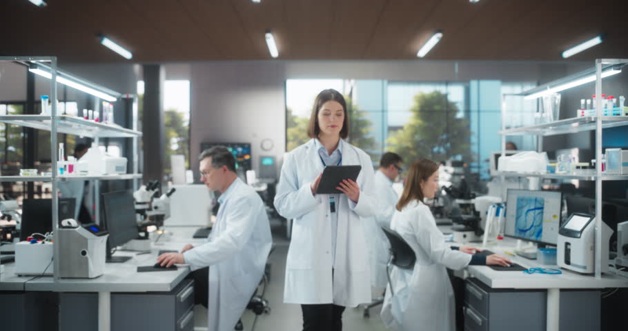 Portrait of an Attractive Female Scientist Posing for Camera, Smiling. Young Biologist Standing in a White Coat, Using a Tablet Computer in a Modern Laboratory, Researching Pharmaceutical Products
