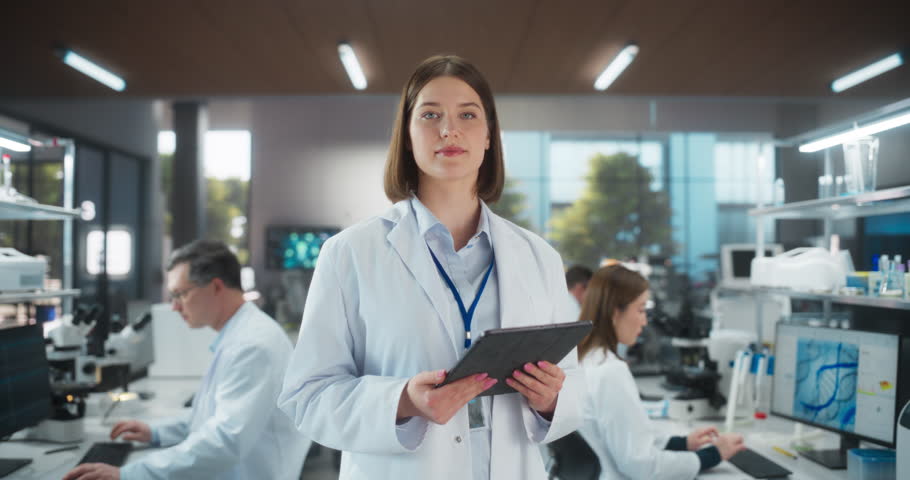 Portrait of an Attractive Female Scientist Posing for Camera, Smiling. Young Biologist Standing in a White Coat, Using a Tablet Computer in a Modern Laboratory, Researching Pharmaceutical Products