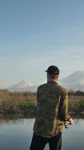 A young adult fisherman in a cap uses a spinning rod with a fishing reel to catch fish in the river against the backdrop of beautiful snow-capped mountains. Catching predatory fish at sunset