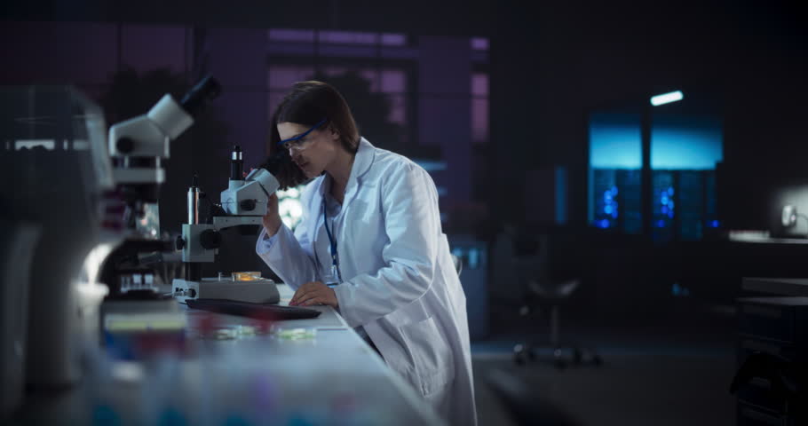Medical Research and Development Laboratory: Hard-Working Female Scientist Using Computer and Looking at a Sample in a Petri Dish Under a Microscope. Advanced Genetic Engineering Lab at Night