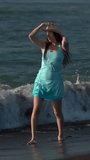 Playful and happiness barefoot woman standing in breaking waves on beach. Full length Caucasian middle aged adult female in straw sun hat, short turquoise summer dress. Handheld. Vertical slow motion