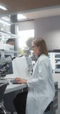 Vertical Screen: Medical Research Scientist Looking at Biological Samples Under a Microscope in a Science Laboratory. Portrait of a Caucasian Lab Engineer in White Coat Looking at Camera and Smiling
