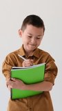 Asian Boy Smiling And Writing Homework In Notebook On White Background