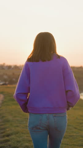 Woman rests walking at sunset riverside. Relaxed slim lady spends tranquil evening on hilly bank of large river at spring dusk. Rural site travel