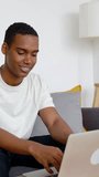 Happy young adult African man using laptop sitting on couch at apartment. Technology and education concept. Vertical HD screen video.