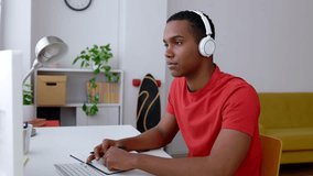 Happy young student boy wearing headphones studying at home. Millennial black male talking on video call while learning or listening online course. Education lifestyle and e-learning concept.