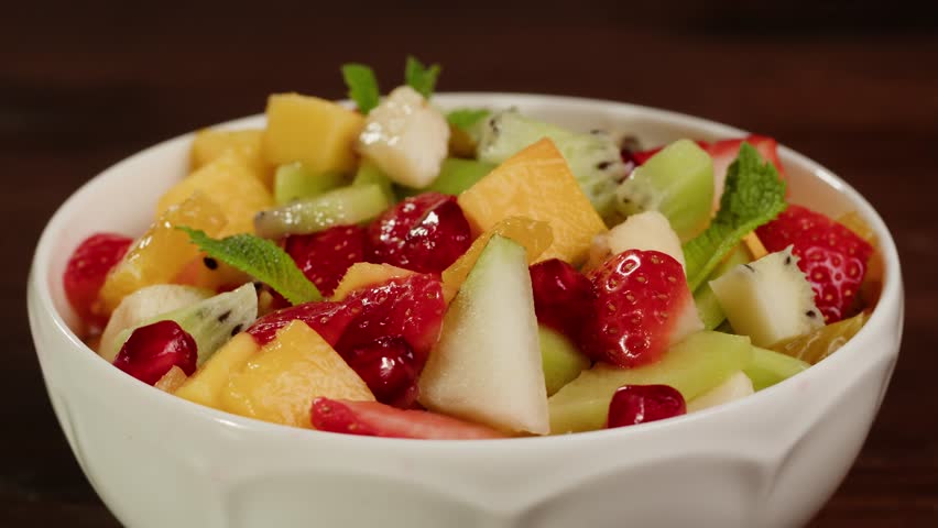Health fresh colorful fruit salad with kiwi, orange, strawberries, mango, banana and mint on the top in bowl on table close-up. Healthy vegan detox summer recipe. Food concept