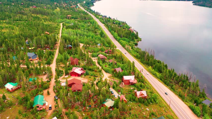 Aerial Drone View of Cars Driving on Road Between Beautiful Clear Blue Lake Water and Forest with Houses and Boat Docks in Grand Lake Colorado During Summer in the Rocky Mountains