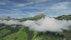 Drone flies smoothly forwards, framing Sass de Putia in cloud-kissed Dolomites. Chalets dot lush hills as drone drifts above them, capturing serene village. Near La Val, Italy. LuPa Creative.