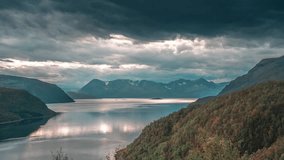 Rays of the setting sun shine through stormy clouds over a calm fjord in a time-lapse video.