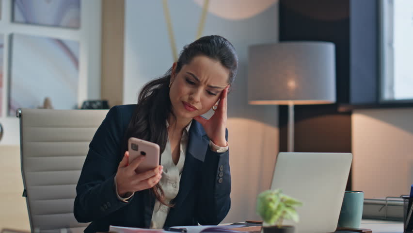 Nervous businesswoman checking smartphone message working computer at office workplace closeup. Tired exhausted woman looking distance worried problems at laptop table. Anxious lady working hard task