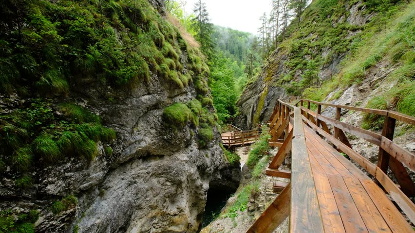 A mountain gorge with a running water creek, surrounded by greenery and rocks. a wooden path caresses the rocks around it, and an unidentified woman is standing in the distance. a 4K video clip.