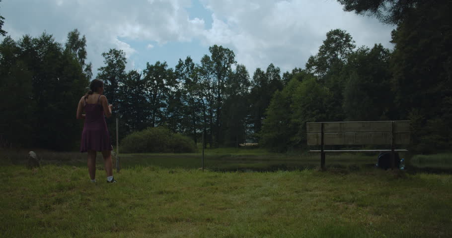 Young woman in dress stands by bench near pond in forest. Relaxing summer life