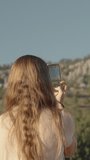 Vertical video. A young woman captures beautiful views on her phone for social media, with tall mountains and trees in the background.
