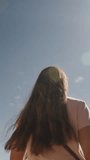 Vertical video. The traveling young woman among ancient ruins and rocks gathers her hair with her hand, against the backdrop of the sun and tall mountains.