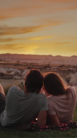 Vertical video. A loving couple, a man and a woman, lie on a cushion in the mountains and watch the sunset, with mountains and a canyon in front of them.