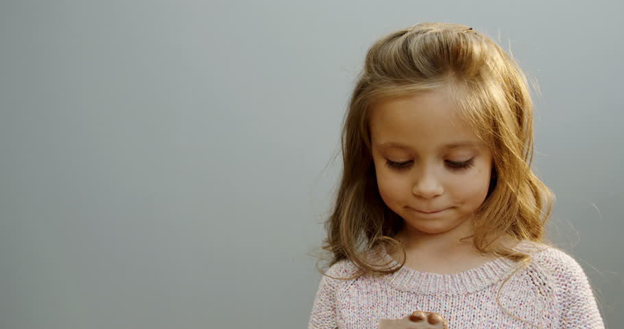 Portrait shot of the cute curly little girl standing in front of the camera and biting a chocolate bar with nuts on the white wall background. Close up. Inside