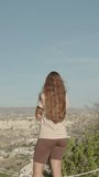 Vertical video. A young woman stands at the top and admires the natural ancient park of Goreme from a high mountain, the camera zooms in on her.