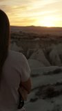Vertical video. View from behind a young woman with long hair, the sun is almost setting behind the distant mountain, and she is enjoying the sunset.