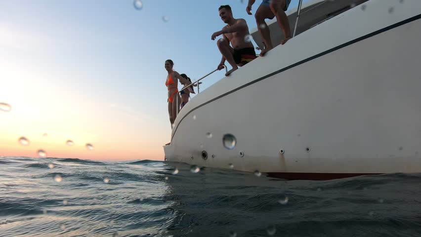 Group of young friends jumping to the beach, having a party in yacht. Attractive man and woman people hanging out, celebrating holiday vacation trip while catamaran boat sailing during summer sunset.